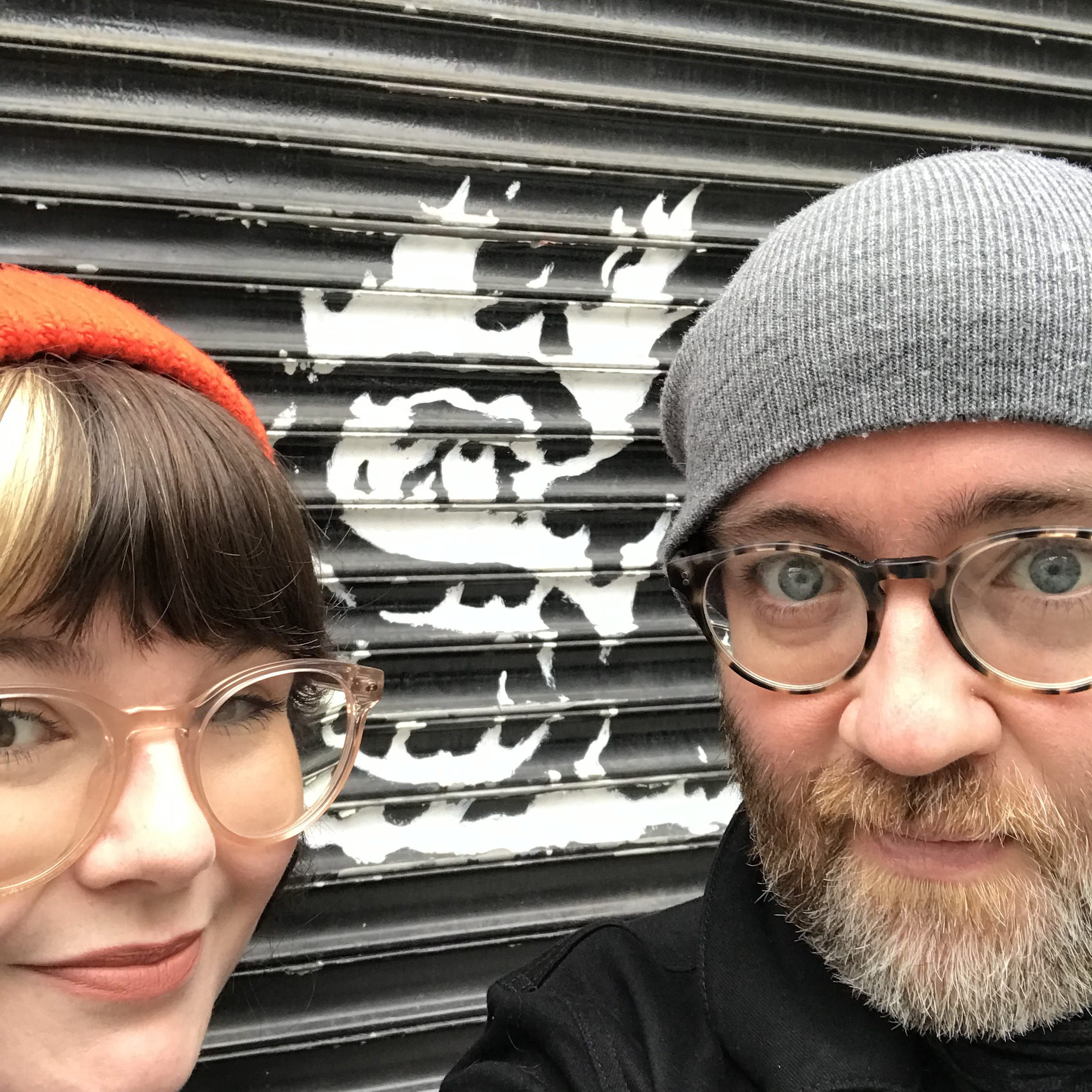 Photograph of bodega shutters, painted black with flaming S logo, with a white women wearing glasses and a white man wearing glasses. 